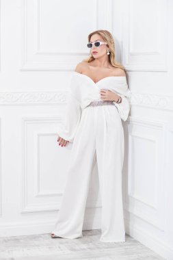 Zengin, orta yaşlı, şık takım elbiseli ve takılı bir kadın klasik beyaz bir iç mekanda poz veriyor. Lüks bir yaşam tarzı. Güzellik, moda.