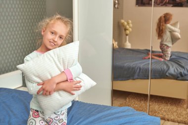 Sevimli küçük kız yastıkla yatakta oturuyor ve kameraya gülümsüyor. Mutlu bir çocukluk. Aile evde..