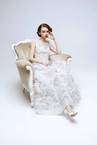 Portrait of a beautiful chic woman posing in a luxury white dress in vintage armchair. Evening makeup and hairstyle of the 20s.