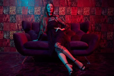 Siyah gece elbisesi içinde güzel bir kadın lüks dairelerdeki bir kanepede oturuyor. Göz kamaştırıcı bir yaşam tarzı. Güzellik, moda. Modern iç mekan, mobilya.