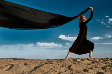 Çölde poz veren uzun siyah elbiseli güzel esmer kadın rüzgarda çırpınan bir eşarpla. Moda fotoğrafı. Gökyüzü arkaplanı.