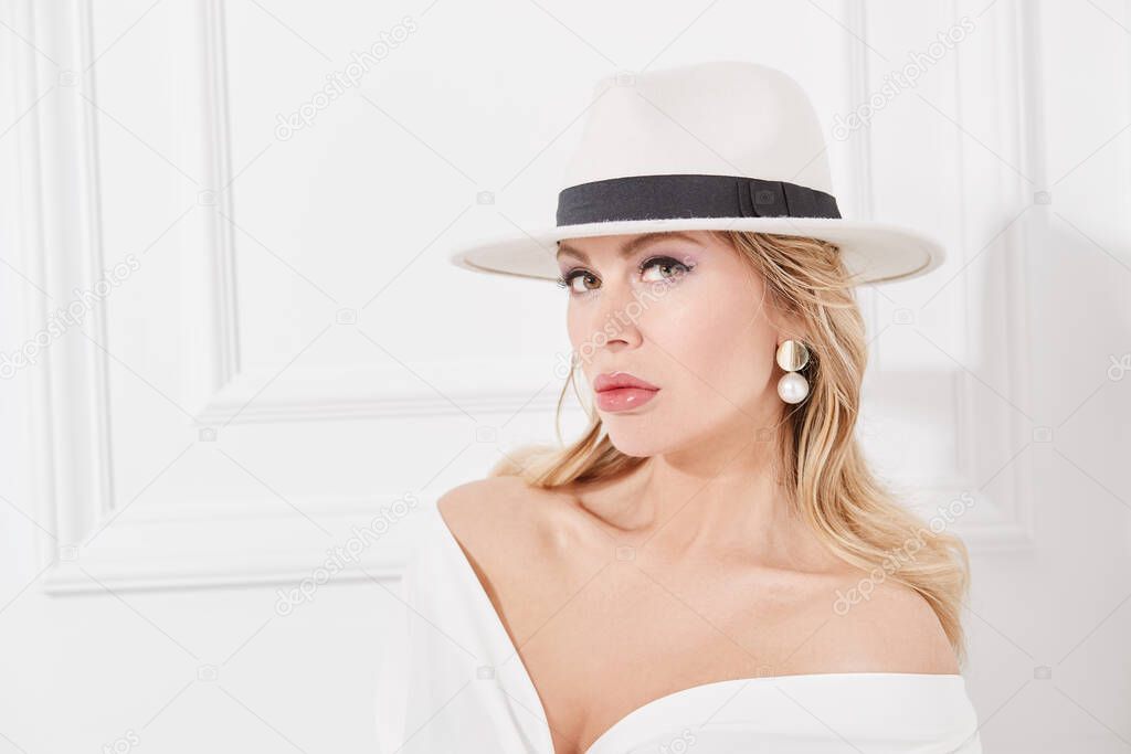 A wealthy middle-aged woman in a chic white suit, elegant hat and jewelry poses in a classic white interior. Luxury lifestyle. Beauty, fashion.