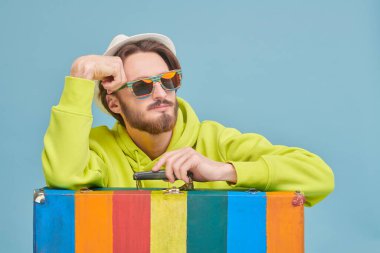 Tatil ve seyahat konsepti. Parlak kapüşonlu, güneş gözlüklü ve parlak renkli bavuluna yaslanmış yaz şapkalı genç bir adam. Mavi arkaplanda stüdyo resmi.