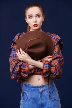 Moda fotoğrafı. Kot pantolon, kareli bluz ve kahverengi şapkayla poz veren güzel bir kız modeli portresi. Country, batı tarzı. Yeni yaz koleksiyonu.