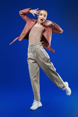 Heyecanlı, pantolonlu, deri ceketli, geniş bir adım atıp koyu mavi arka planda ağzını açan modern bir kız modelin portresi. Güzellik, moda. 