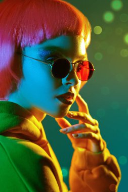 Parlak makyajlı, pembe saçlı, kırmızı güneş gözlüklü, sarı kapüşonlu, karışık renkli ışık saçan modern bir kız. Sportif şıklıkta. Gençlik modası ve güzellik eğilimleri. 