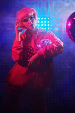 Parlak makyajlı, pembe saçlı çekici kız disko topu ve neon ışıklı kulaklıklarla poz veriyor. Disko kızı. Gece partisi. DJ kız. 