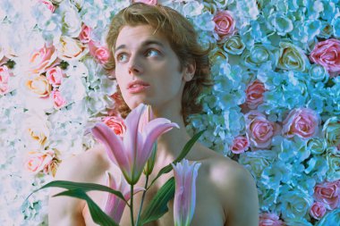 Parfüm ve güzellik ürünleri. Sarı dalgalı saçlı, pembe beyaz zambaklar ve güllerle poz veren romantik genç bir adam. Erkeklerin güzelliği. 