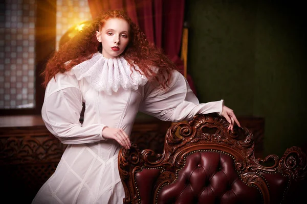 History of fashion and hairstyles. A stylish fashion model girl with lush red hair with fine curls poses by a vintage armchair in art dress with a ruffled renaissance collar.