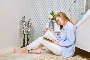 Güzel kız rahat, parlak yatak odasında dinleniyor, yatağın yanında oturuyor ve bir kitap okuyor. Evin içi, mobilyalar. Yaşam biçimi.