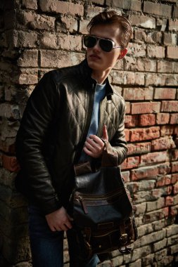 Güneş gözlüklü, deri ceketli, kendine güvenen ve şık genç bir adam şehrin bir sokağında tuğla bir duvarın yanında duruyor. Erkek modası. Yaşam biçimi.