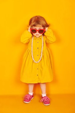 Parlak sarı bir arka planda kırmızı gözlüklü, boncuklu ve sarı elbiseli şirin küçük bir kızın tam boy portresi. Çocukların yaz modası.. 