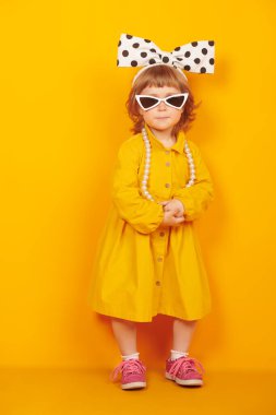 Parlak sarı elbiseli, büyük yay ve sarı arka planda duran güneş gözlüklü güzel bir kızın tam boy portresi. Pin-up stili. Çocuk modası.. 