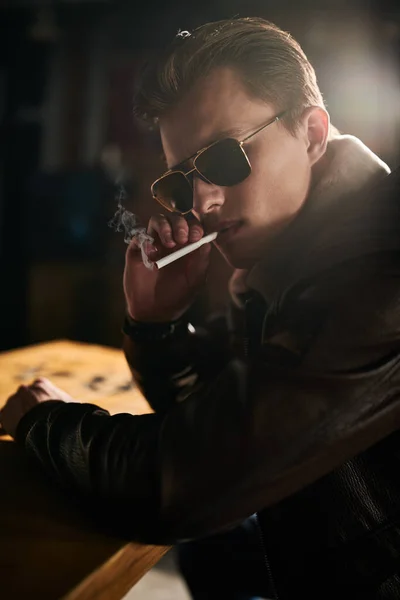 Handsome young man in a leather jacket and sunglasses sits at the bar and smokes a cigarette. Lifestyle.
