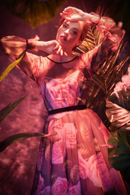 Çiçekler ve tropik yeşillikler arasında poz veren açık renkli çiçekli genç bir kadın. Yaz güzelliği, moda. 