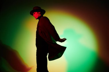 Sanat portresi. Zarif şapkalı ve ceketli yakışıklı genç adam stüdyoda karışık renkli ışıkta poz veriyor. Erkek güzelliği, moda.