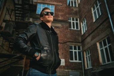 Siyah deri ceketli ve siyah güneş gözlüklü yakışıklı adam şehrin eski semtinde caddede duruyor. Kentsel grunge tarzı. 