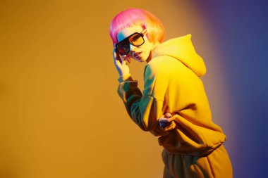 Parlak pembe saçlı modern kız sarı güneş gözlüklü ve sarı takım elbiseli karışık renkli ışıkta poz veriyor. Sportif şıklıkta. Gençlik modası ve güzellik eğilimleri. 