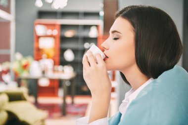 Mutlu genç bir kadın oturur, battaniyeye sarılır ve kahve içer. Güzel, mutlu kız, rahat, aydınlık salonunda dinleniyor. Mutlu insanlar ve yaşam tarzı konsepti.