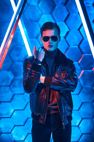 Мужской стиль и мода. Портрет мужественного красавца в черных солнцезащитных очках и черной кожаной куртке, позирующей среди неоновых ламп. Футуризм, техно стиль.