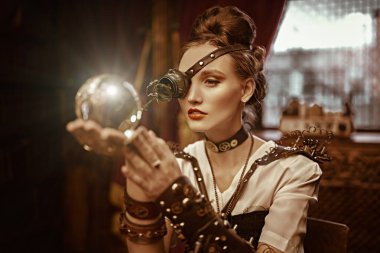 Steampunk 'ın gizemli dünyası. Güzel steampunk kadın bilim adamı, Viktorya dönemi iç mimarisi ile laboratuarında duran bir kristal küre tutuyor.. 