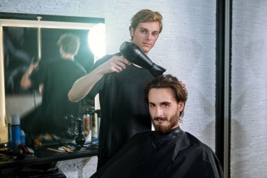 Erkek güzeli. Usta berber saç stilini ve saç modelini kurutma makinesiyle yapıyor, yakışıklı bir erkek müşteriye saç kurutuyor. Berber Dükkanı.