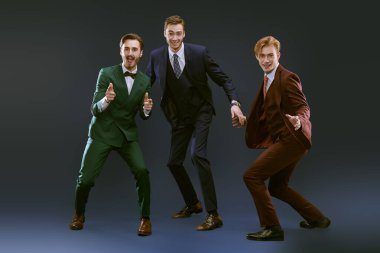 Zarif takım elbiseli üç yakışıklı erkek bir arada durup gülüyor. Üç arkadaş. Erkek modası. Koyu mavi arkaplan.