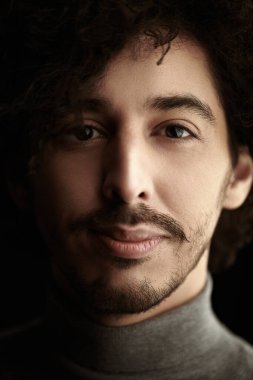 Kıvırcık siyah saçlı, kameraya bakıp gülümseyen yakışıklı bir gencin portresi. Siyah arka plan. Erkek sağlığı ve güzelliği.