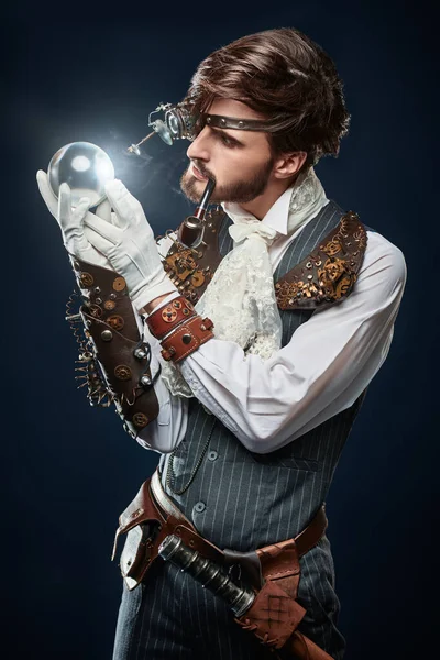 Fantasy world. Portrait of a handsome steampunk man looking at the crystal ball on a dark blue background.