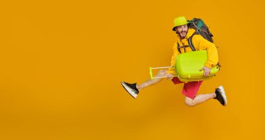 Sırt çantası ve bavullarla atlayan duygusal genç hippi portresi. Turizm, seyahat, tatil. Fotokopi alanı olan sarı bir arkaplanda stüdyo resmi.