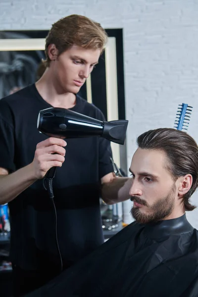 Male beauty. Master barber does the hairstyle and styling with dryer, dries hair to a handsome male client. Barbershop.