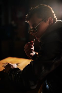 Deri ceketli ve güneş gözlüklü yakışıklı genç adam barda oturur ve karanlıkta sigara içer. Yaşam biçimi. 
