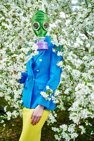 A woman in blue jacket and green gas mask stands in a blossoming apple orchard on a clear spring day. Air pollution, environmental disaster.