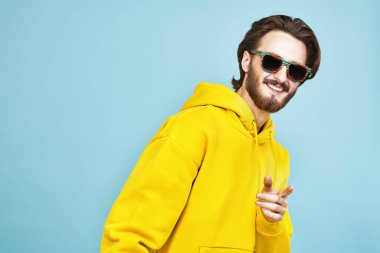 Güneş gözlüklü ve parlak kapüşonlu genç adam kameraya parmağını gösteriyor. İnsanlar, duygular. Fotokopi alanı olan mavi bir arkaplanda stüdyo resmi.