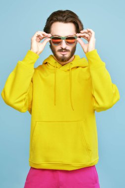 Güneş gözlüklü ve parlak kapüşonlu genç adam stüdyoda mavi arka planda poz veriyor. Gençlik modası. Optikler.