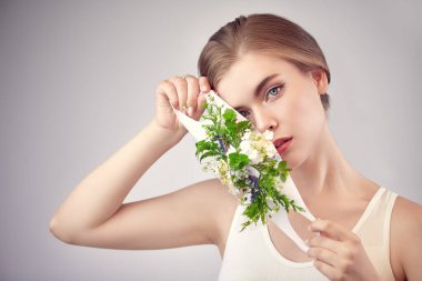 Açık gri arka planda yeşil bitkiler ve çiçeklerle süslenmiş güzel sarışın bir kızın portresi. Moda, güzellik ve sağlık hizmetleri. Stüdyo resmi.  