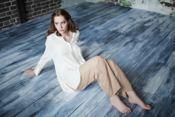 Portrait of a beautiful young woman with light makeup, wavy hair and in light white shirt sitting on the floor in a room with brick walls. Beauty, fashion. People and lifestyle.