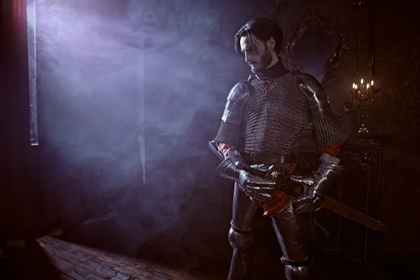 The noble knight in armor knelt down with his sword and looks at the light in the castle. Romance.