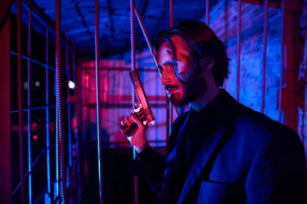 Portrait of a handsome wounded special agent standing on alert with a gun in his hand behind bars in multicolor lighting.