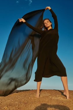 Uçma hissi, rüyalar. Uzun siyah elbiseli güzel romantik bir kız çölde poz veriyor rüzgarda çırpınan bir eşarpla. Gökyüzü arkaplanı.
