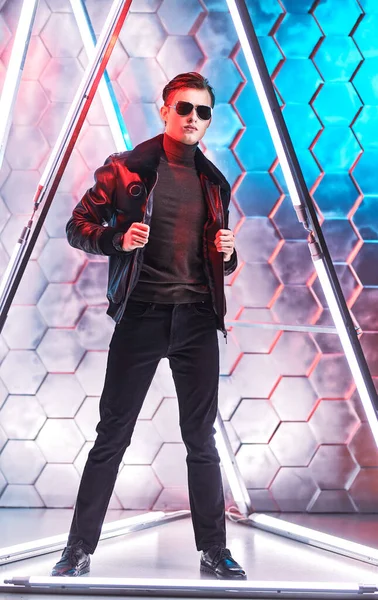 Men\'s style and fashion. Full length portrait of a courageous handsome man in black sunglasses and black leather jacket posing among the neon lamps. Futurism, techno style.