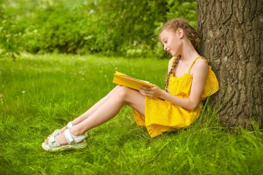 Saçları örgülü, gülümseyen tatlı bir kız elinde kitapla yeşil bir çimenlikte oturuyor. Mutlu yaz çocukları. Parkta yürü..