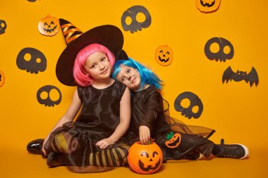 Çocukların cadılar bayramı. Cadı kostümü giymiş iki tatlı kız ve bir sepet şekerle siyah kedi şakası ya da şeker. Sarı arka planda stüdyo resmi. 