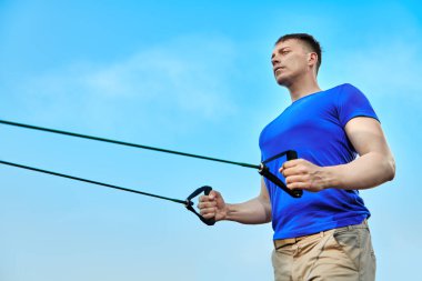 Açık spor salonunda çalışıyorum. Atletik bir adam mavi gökyüzüne karşı elastik bantlarla güç egzersizleri yapar. Aktif sağlıklı yaşam tarzı. 