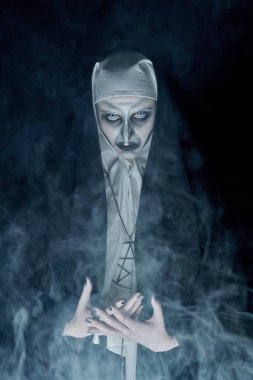 Cadılar Bayramı ve dehşetler. Gizemli bir dumanla siyah arka planda kameraya bakan korkunç şeytani bir rahibenin portresi.. 