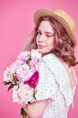 Açık beyaz yaz elbisesi ve hasır şapkalı, pembe arka planda şakayık demeti olan güzel bir kız. Yaz güzelliği ve moda. Mutlu insanlar..