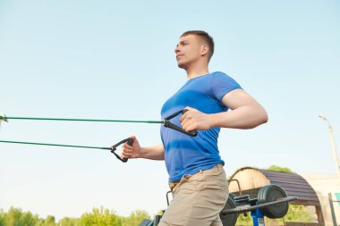 Açık spor salonunda çalışıyorum. Yakışıklı kaslı adam dışarıda spor salonunda elastik bantlarla güç egzersizleri yapıyor. Aktif sağlıklı yaşam tarzı. 