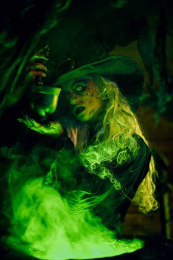 Korkunç yaşlı cadı, sihirli yeşil ışıkla çevrili kazanında iksir yapıyor. Cadılar Bayramı. Korkunç hikayeler..