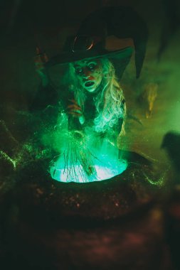 Korkunç yaşlı cadı, sihirli yeşil ışıkla çevrili bir kazanda kaynayan iksirin üzerinde süpürge tutuyor. Cadılar Bayramı. Büyücülük..