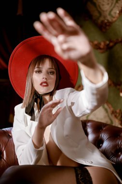 Tutkulu, üstsüz, beyaz ceketli esmer bir kız, siyah dantelli çoraplar ve klasik bir odada deri koltukta duran zarif kırmızı şapkalı bir kız.. 